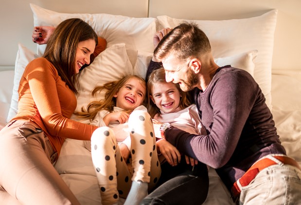 Happy family laying and resting on the bed with their kids, two daughters, in a hotel room. Mother, father and daughters cuddling and tickling on the bed in a hotel room.