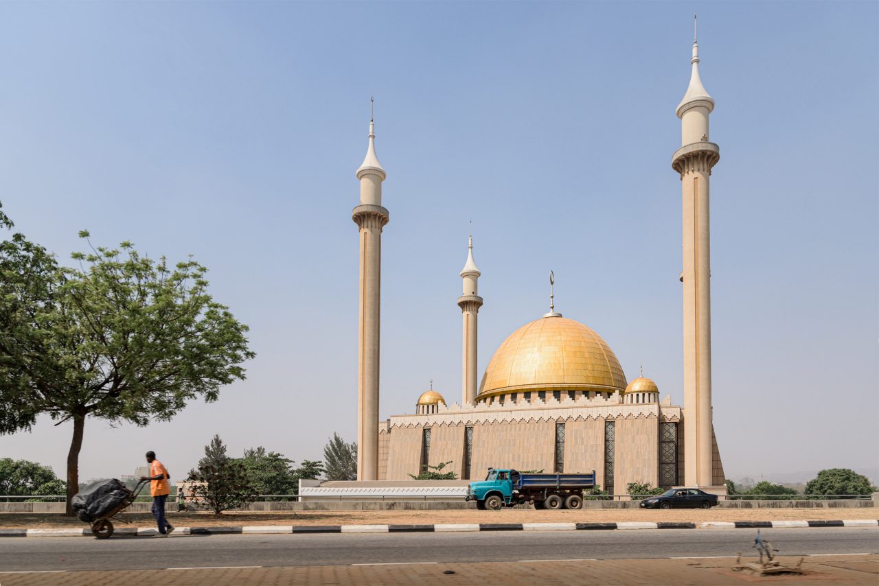 Abuja, Nigeria - 27 Jan 2021: A man pushes a cartwheel along an empty highway in front of the National Mosque landmark building in Abuja, Nigeria. The West Africa city sees no street traffic following Covid-19 confinement restrictions.